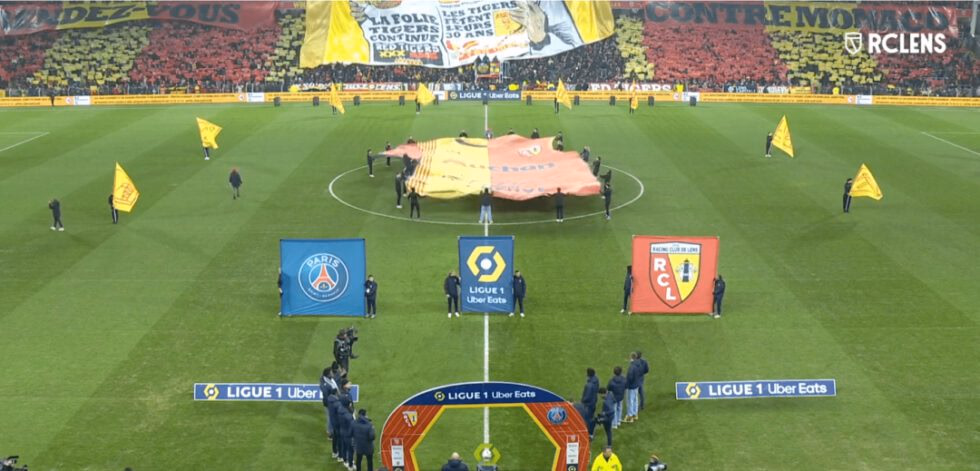 Maillot géant RC Lens déployé au rond central lors d'un match de Ligue 1, avec tifo et drapeaux géants — réalisation Nakara Sport